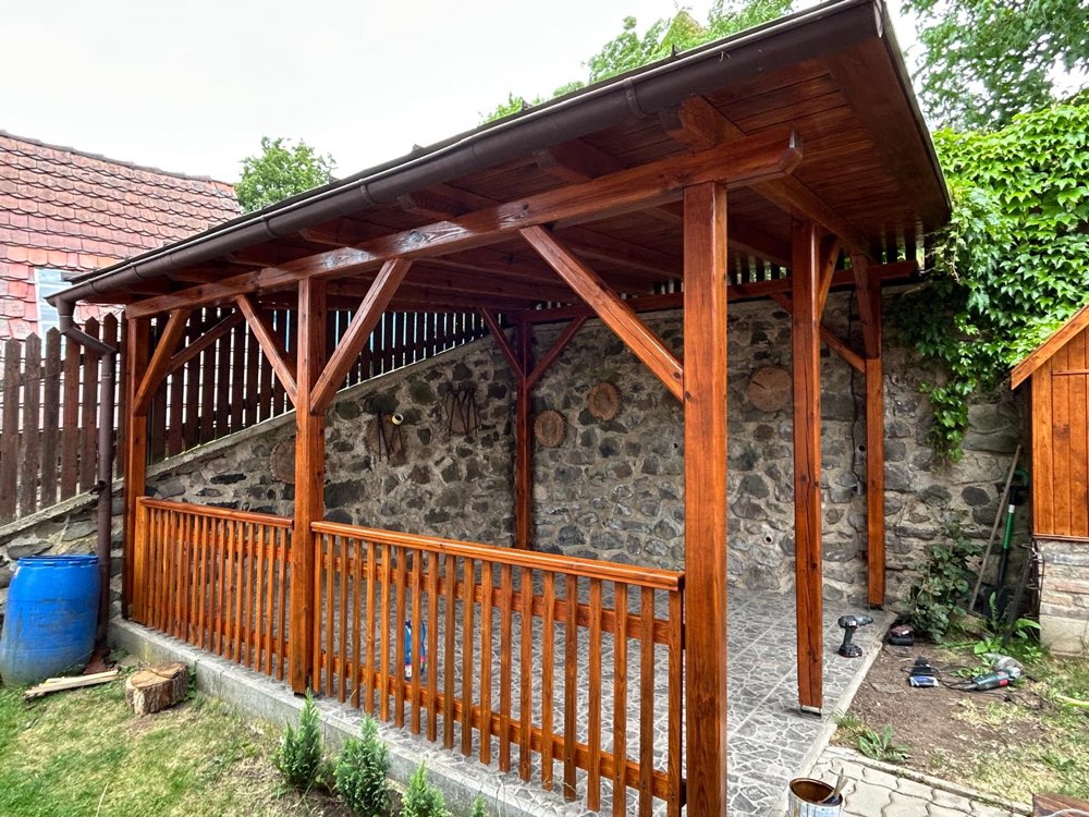 Dřevěná pergola s čerstvým nátěrem v medovém odstínu, opřená o kamennou zeď zahrady.